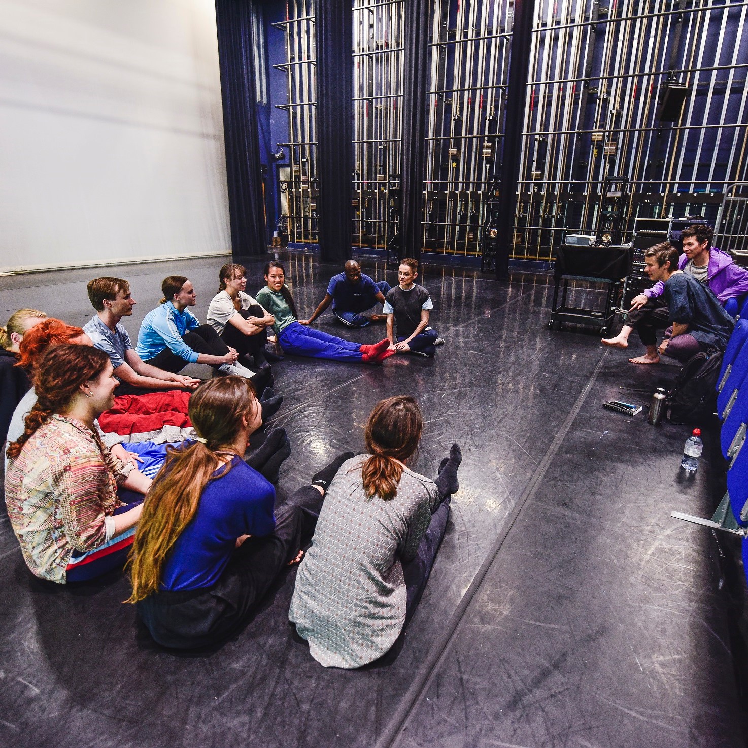 dancer sit in a circle on the floor of the blue room listening to a choreographer talk