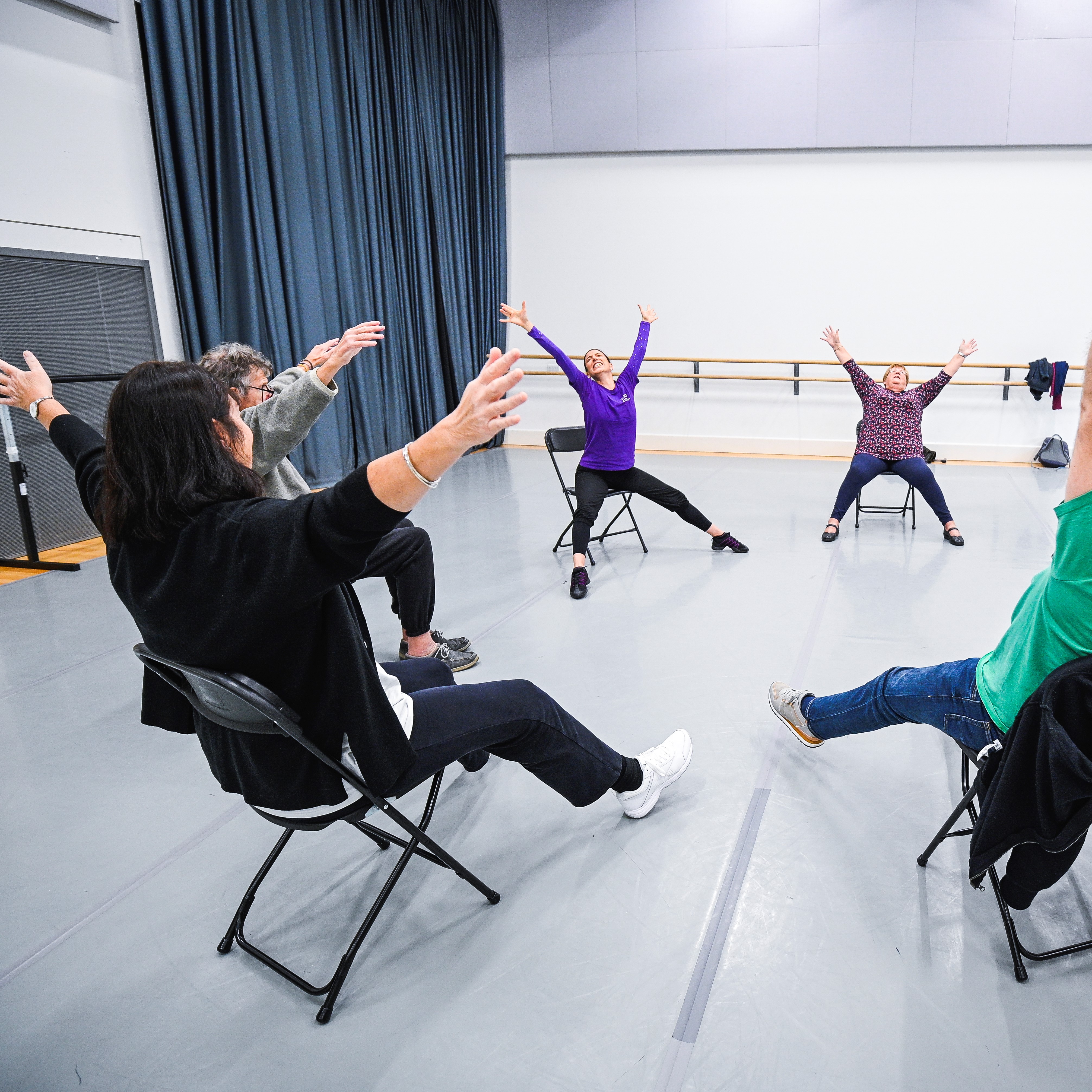 older dancers sit in circle on chairs stretching their arms and legs in a star shape in the man gwyn studio