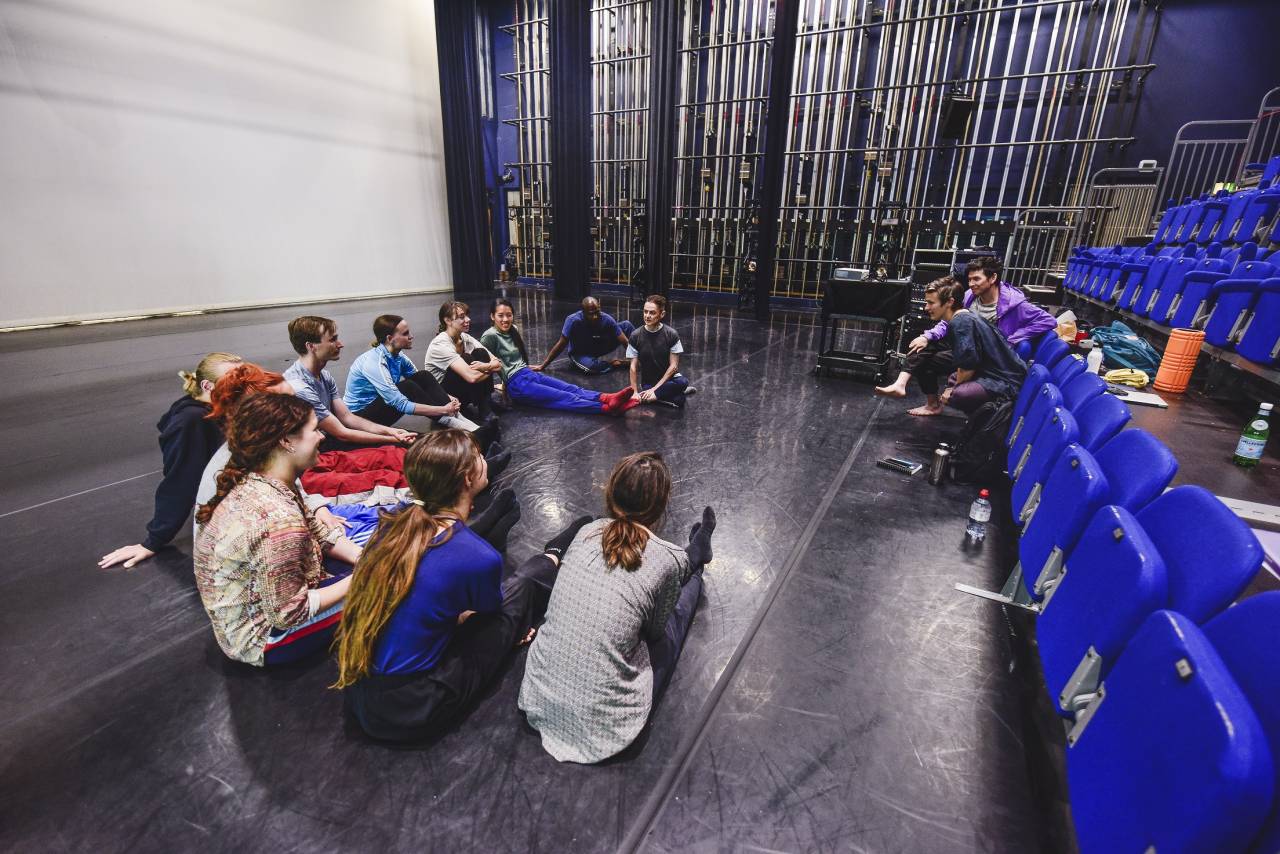 dancers sat on the floor in a circle facing a choreographer sat on blue chairs