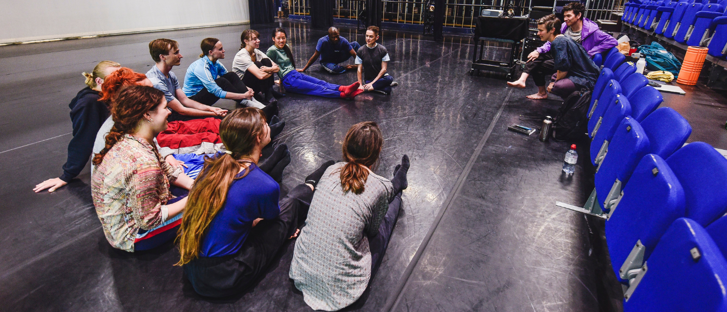 10 dancers in casual clothes sit in a circle avidly discussing something with a male and a famale choreographer - they're in a black floored dance studio
