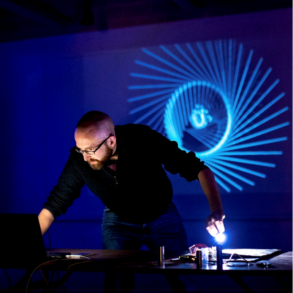 Richard leans over a laptop and dj decks, the back wall behind him lit with projections from his work