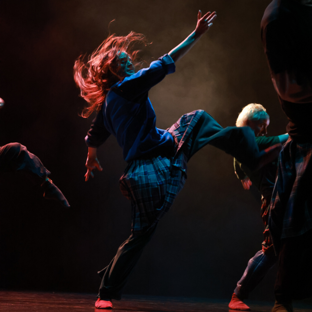 a dancer in a tartan skirt dances under warm dramatic blue and orange lights, long hair flying upwards with momentum and a leg and arm in the air