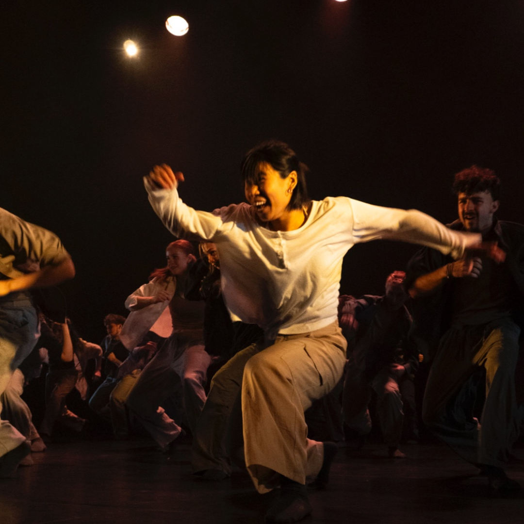 a dancer in tan colour loose clothing swings their arms and grins widely whilst lunging towards the left of the camera