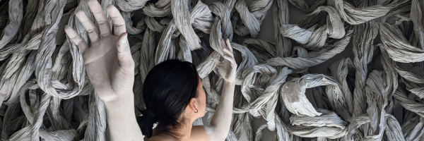 a dancer turns away from the camera holding her hands above her head, her arms are painted stark white, and before her is a large paper sculpture