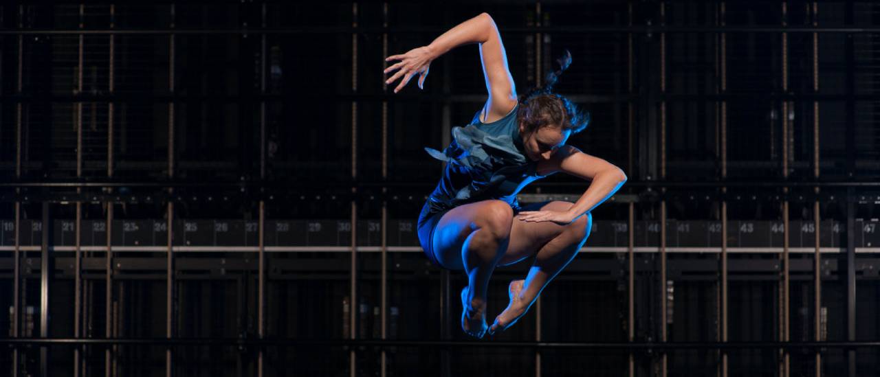 Dancer wearinf a blue dress, jumping in the air legs in a ball with one arm at a 90 degree angle