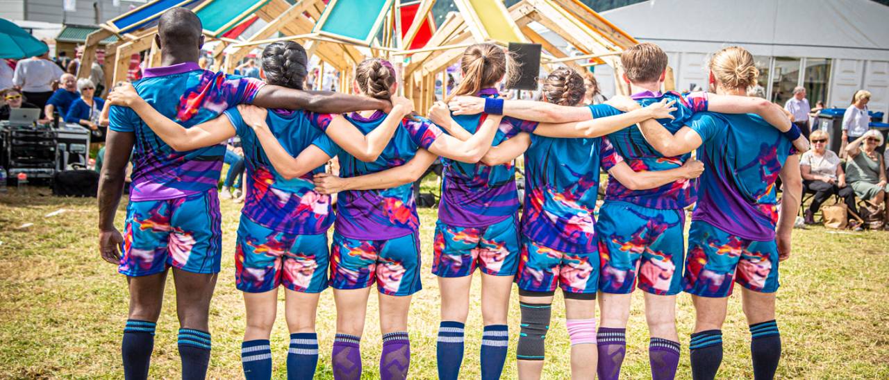 Image of 7 dancers with their backs to the camera, arms linked behind their backs. They are wearing bright coloured sports kits