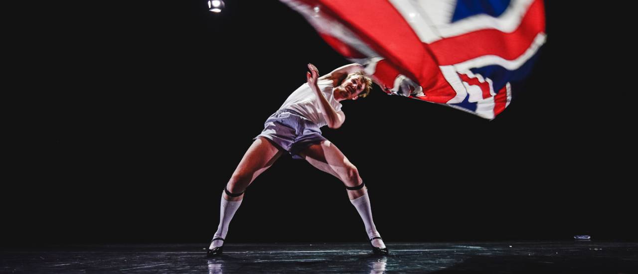 1 male dancer legs spread out, wearing stilettos waving the union jack over his head