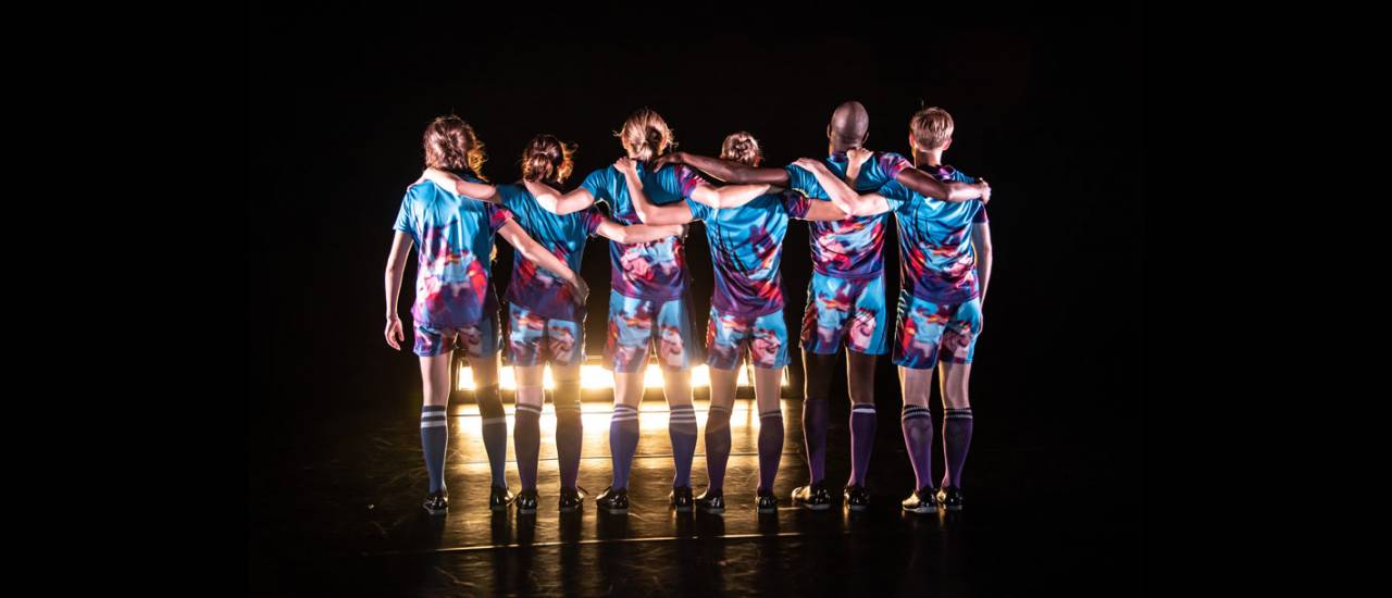 Dancers facing the back of the stage in a line, arms over their shoulders