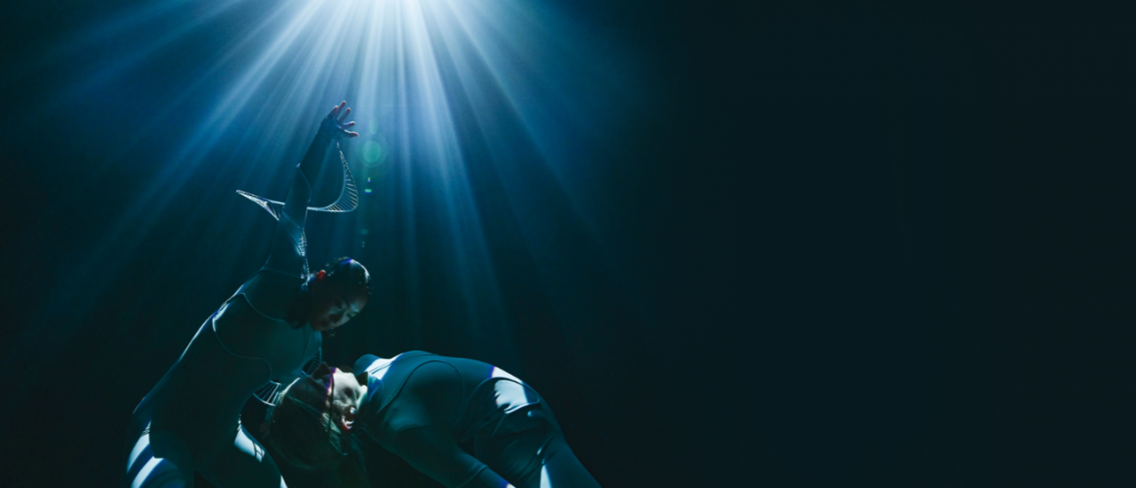 two dancers in futuristic costumes bend backwards under rays of light