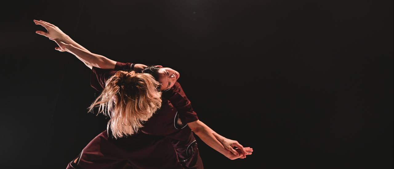 two dancers one leaning over, the other backbending across them to create one body with two heads, arms outstretched