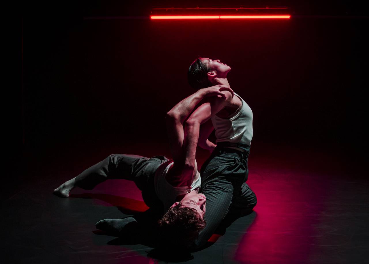 two dancers under a red light, one on the floor the other on their knees above, bending backwards