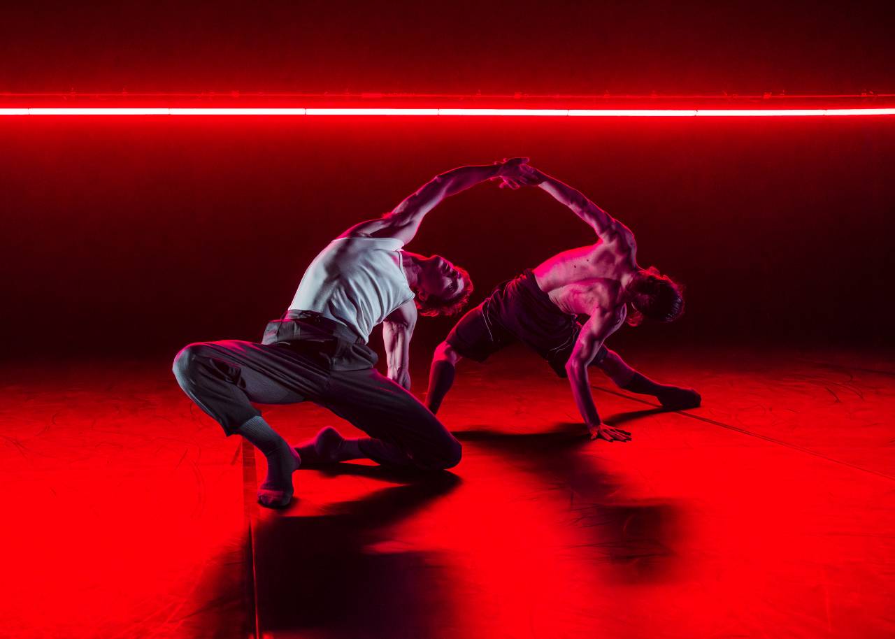 two dancers under red light arch backwards, one hand and two feet splayed across the floor, mid movement