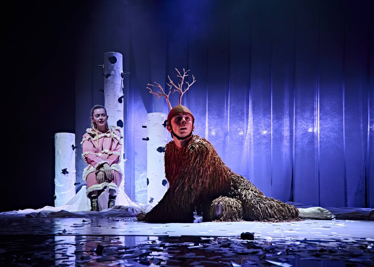 a dancer dressed as a deer sits on all fours on a blue lit wintery stage as a singer sits behind him in a pink winter outfit that looks scandinavian, they both look wistful