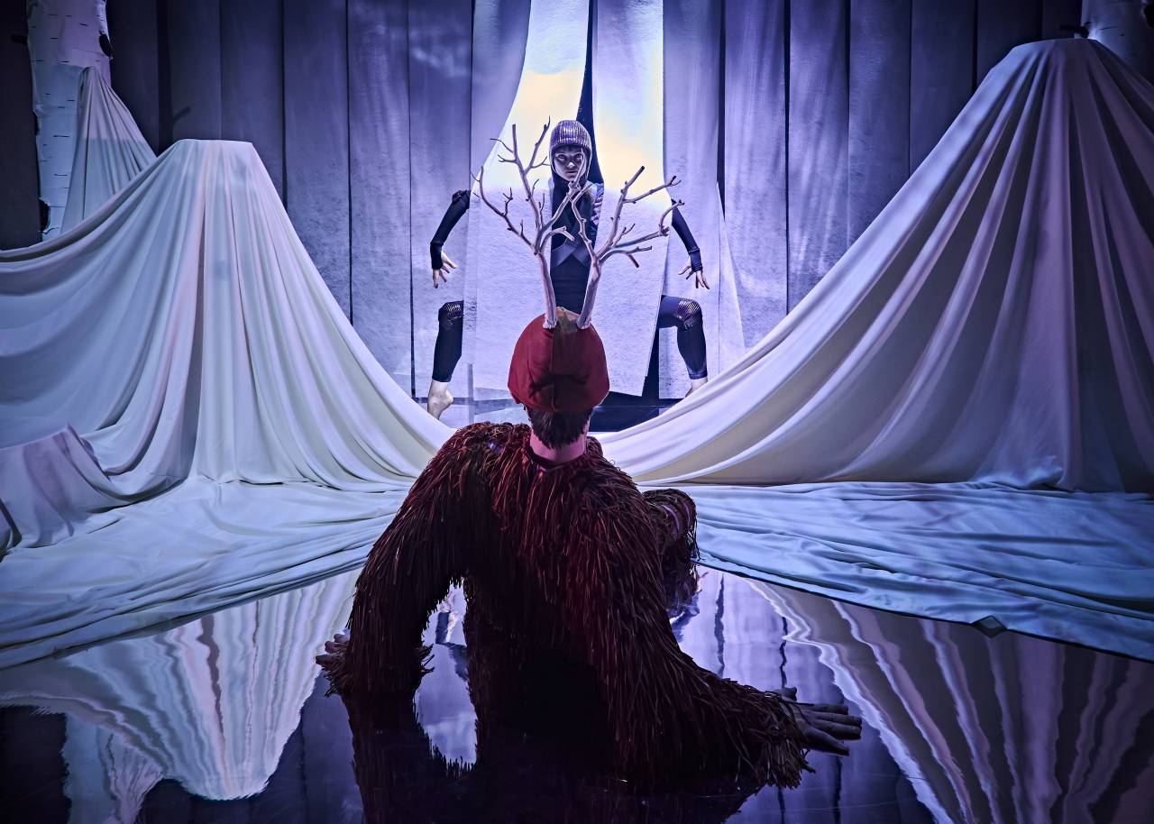 a dancer dressed as a deer leans back, facing away from the camera, they are looking at another dancer dressed as a spider, limbs all bent, hiding in the paper strips that make up the back of the set 