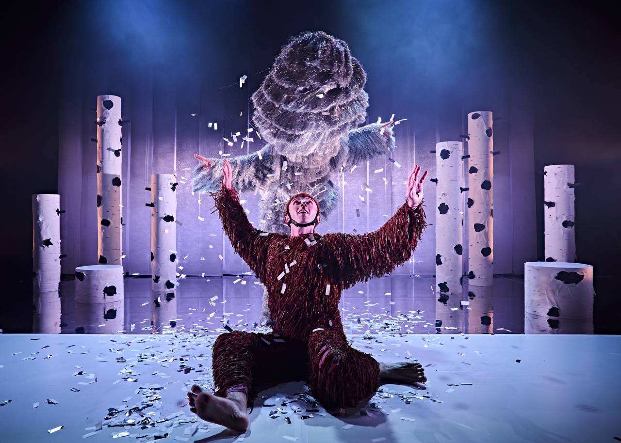 a dancer dressed as a deer sits on the floor arms in the air in joy as another dancer in a huge silver costume made of tassles shakes snow confetti over them - they're in a wintery inspired set with wooden trees