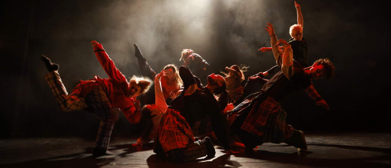 a group of young dancers in cool yellow tartan costumes with arms and legs in the air creating strong shapes under smoke and bright lights