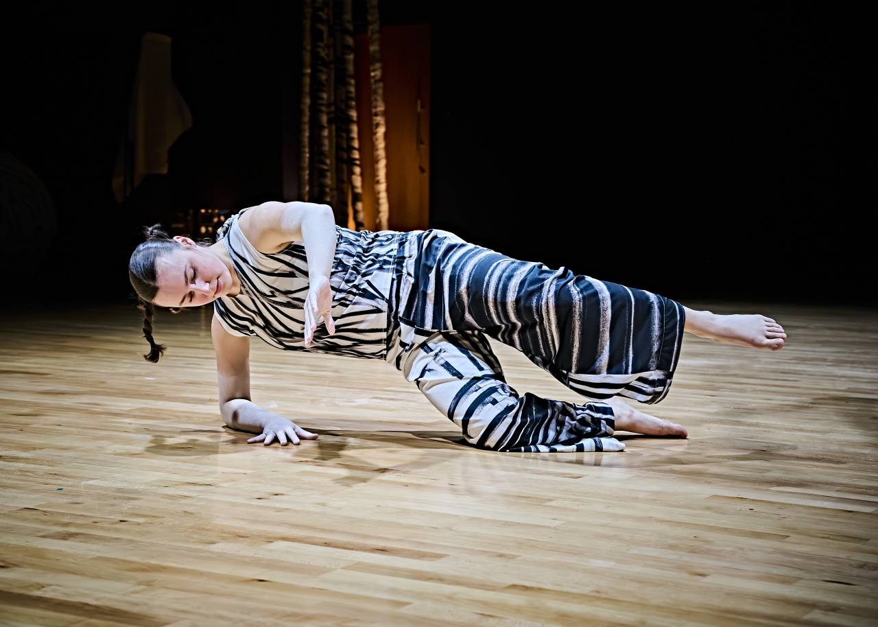 a dancer wears a costume with printed sketches from artist Cecile Johnson Soliz she balances on one knee and one elbow on a wooden floor, seemingly suspended in the moment