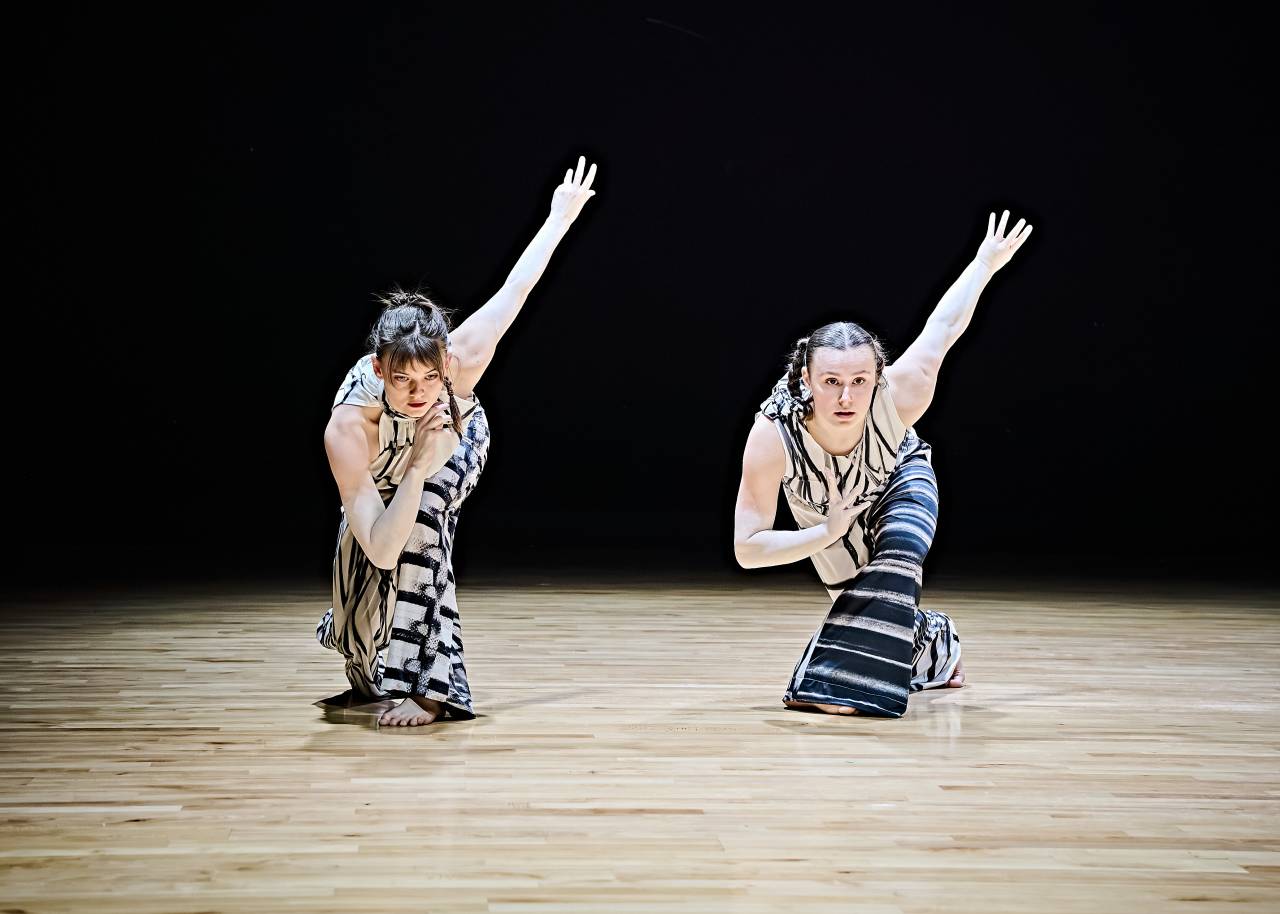 two dancers wearing costume with printed sketches from artist Cecile Johnson Soliz bend forward on one knee, facing the camea, one hand on their heart, the other stretched towards the sky behind them