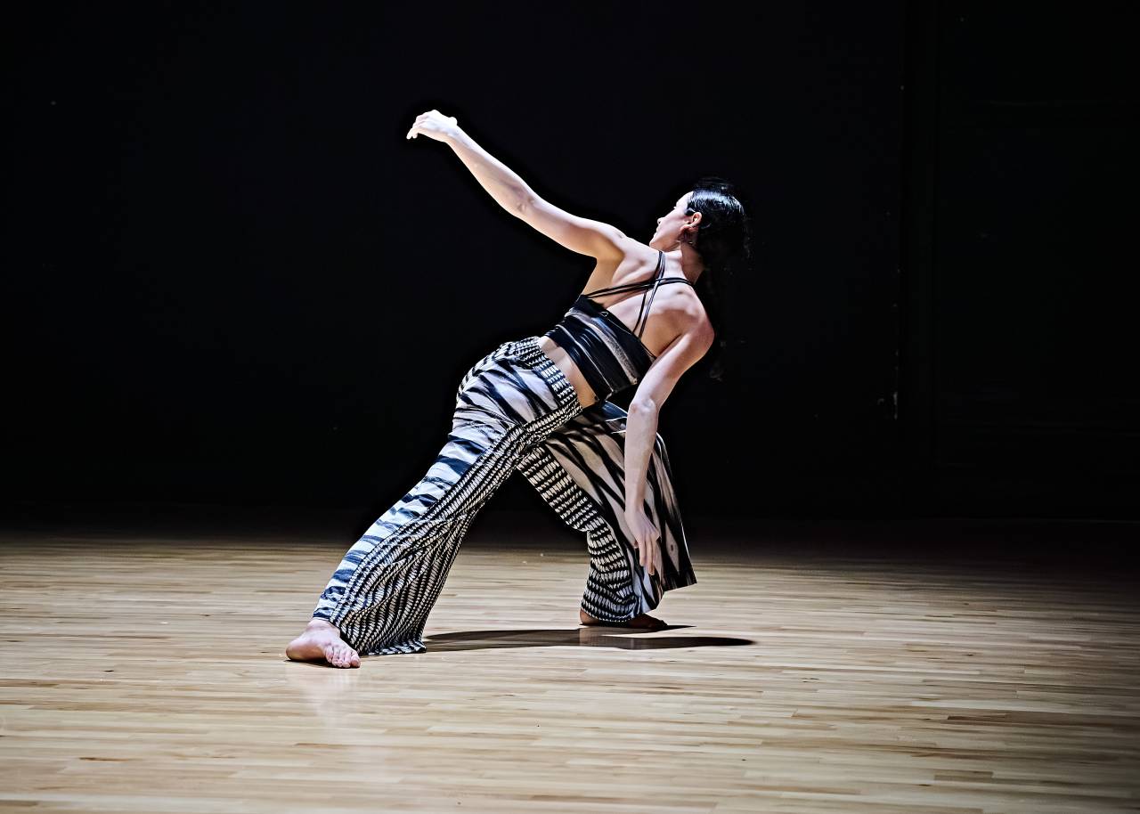 dancer with black hair leans deeply forward, one knee bent the other straight, and arm out behind her, she wears a costume with printed sketches from artist Cecile Johnson Soliz