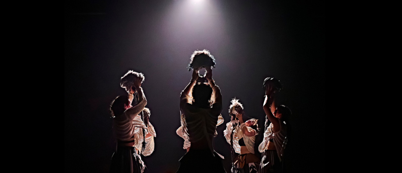dancers in wide, strong stances hold helmets over their heads, stretching up towards a dramatic spotlight 