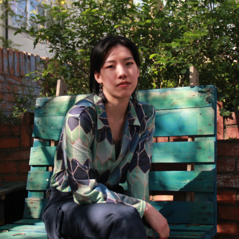 Faye tan sits outside in warm sunlight on a teal bench made of palettes, she has a green pattered blouse on and leans casually on her knee, she has short black hair