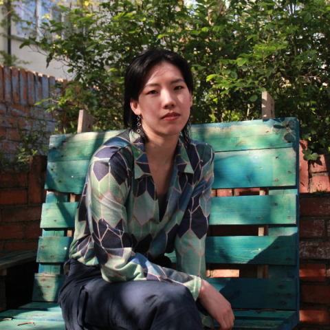 Faye tan sits outside in warm sunlight on a teal bench made of palettes, she has a green pattered blouse on and leans casually on her knee, she has short black hair 