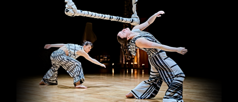 two dancers bend backwards under a large swinging paper sculpture, all mid-movement, they wear white loose costumes printed with black line sketches