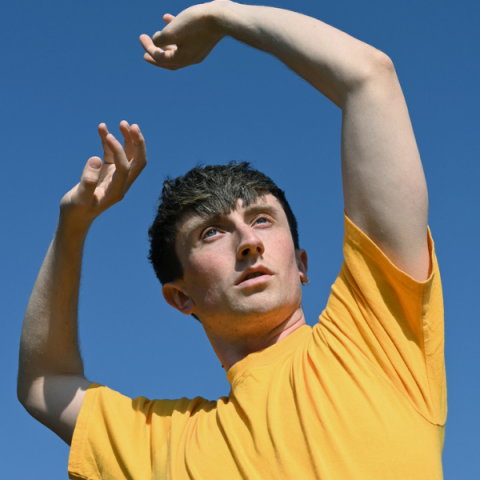 Osian in a yellow t-shirt against a bright blue sky, thier arms up ovre their head